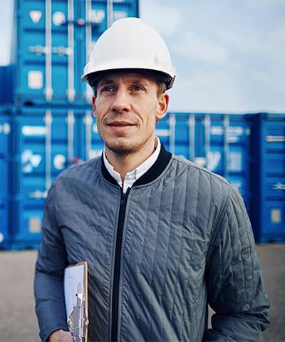 Momento Comenzar a exportar e importar - Trabajador con casco blanco y chaqueta azul sonriendo mientras trabaja en un puerto rodeado de contenedores